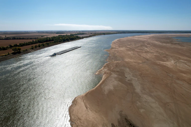 A barge moves through a narrow channel next to a large exposed sandbank