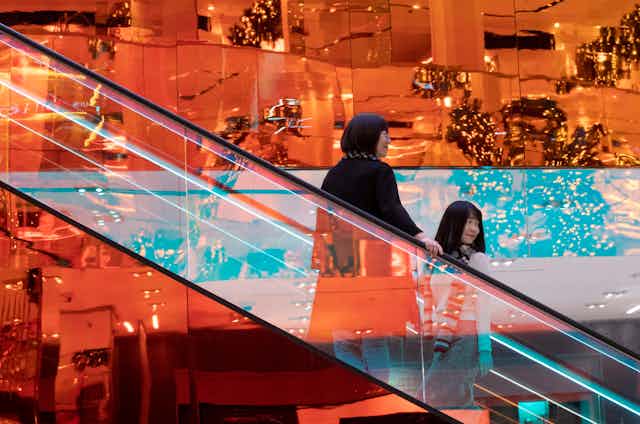 Two women ride a gleaming, golden escalator with Christmas tress in the background.