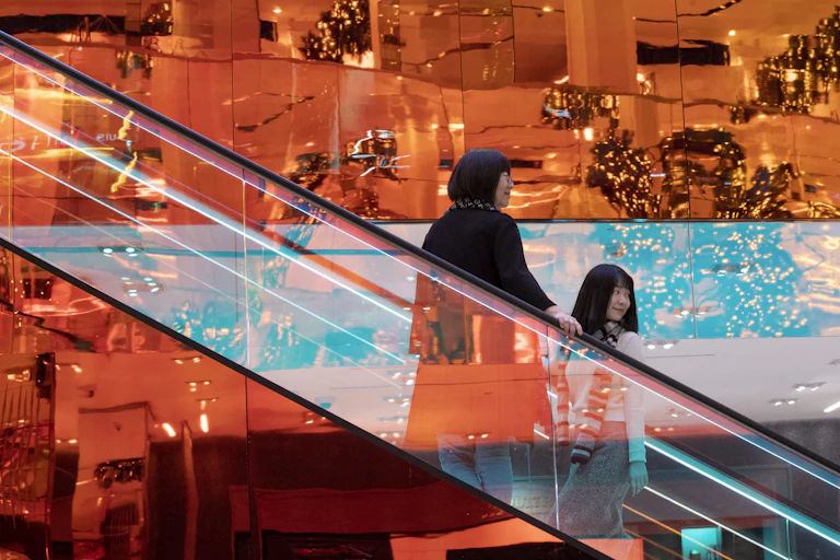Two women ride a gleaming, golden escalator with Christmas tress in the background.