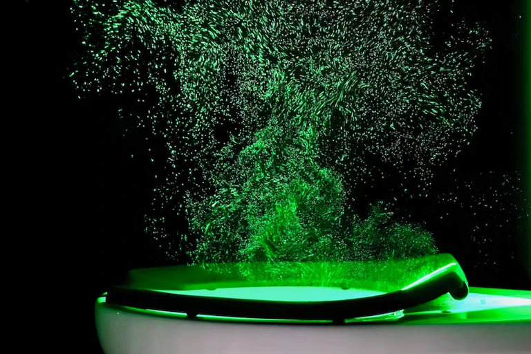 Panache de particules illuminées par un laser au-dessus d'un toilette dont la chasse est tirée