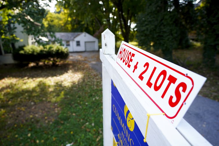 a for sale sign hangs on a white post in front of a home pictured in the distance