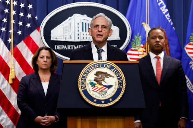 A white haired man standing behind a lectern emblazoned with the seal of the US Department of Justice, speaking.