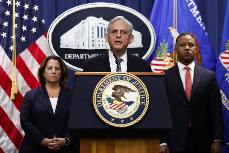 A white haired man standing behind a lectern emblazoned with the seal of the US Department of Justice, speaking.