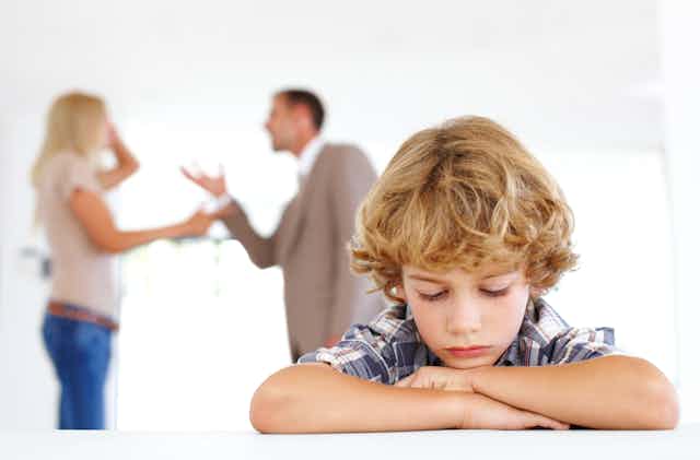 A dejected boy rests his head on his hands as his parents argue in the background.