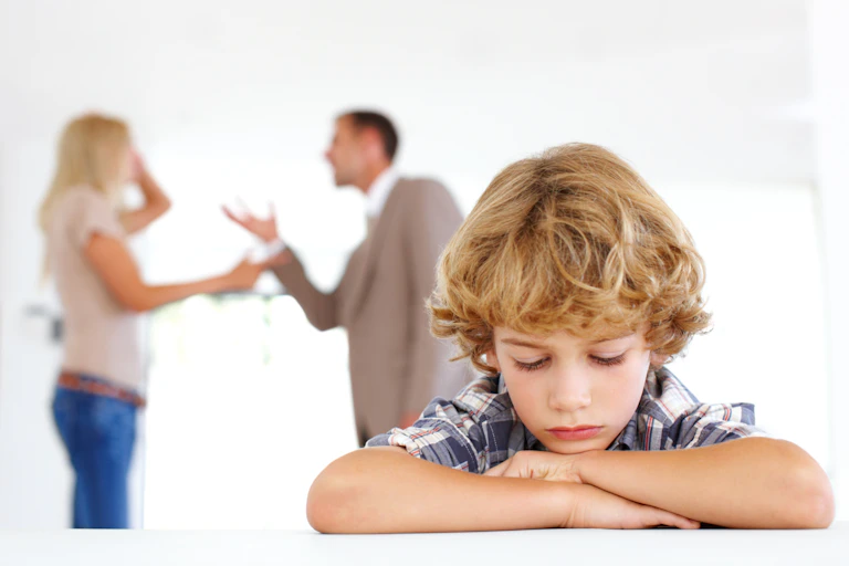 A dejected boy rests his head on his hands as his parents argue in the background.