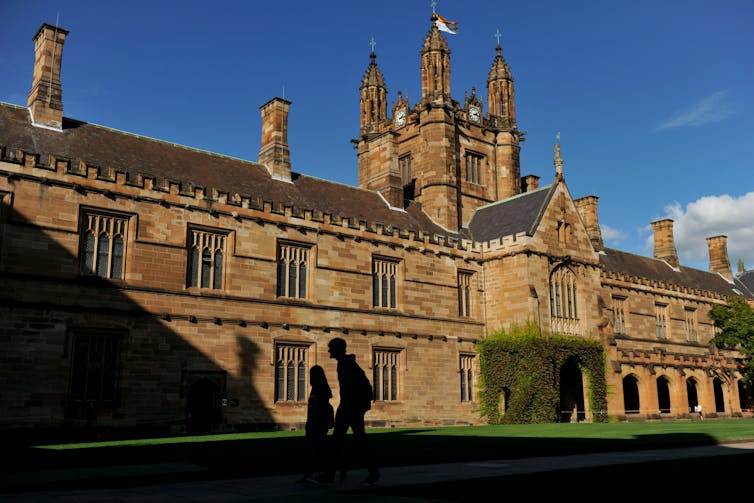The shadows of two students against sandstone buildings at Sydney University.