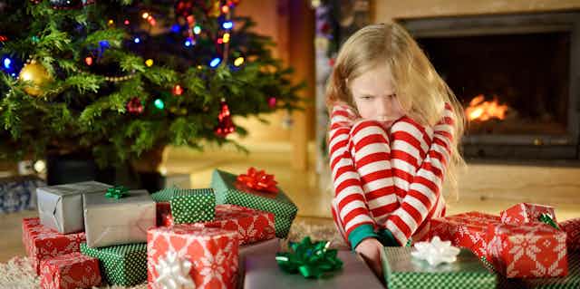Girl child contemplating wrapped gifts besides Christmas tree.  
