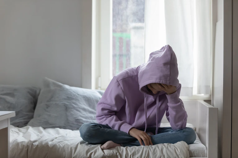 Teen in a purple hoodie sits on their bed