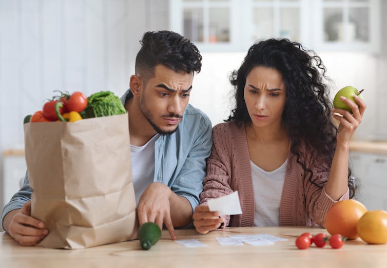 A young couple sits at their kitchen table and looks quizzically at receipts while holding a large bag of fruits and vegetables