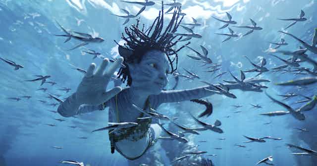 A young n'avi character swims underwater surrounded by fish.
