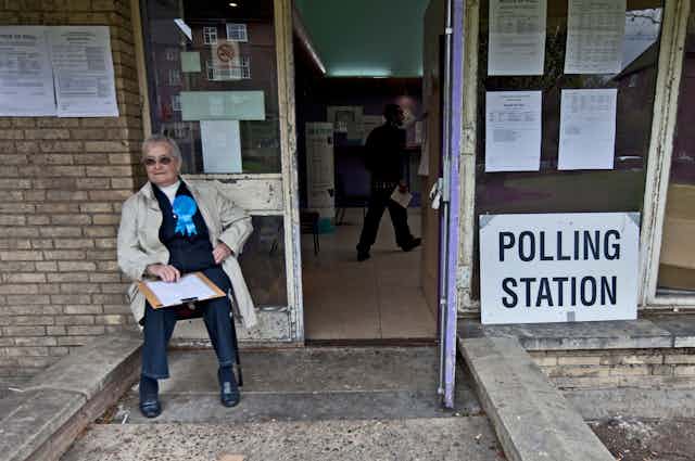 A Conservative party representative sitting on a chair outside a polling station. 