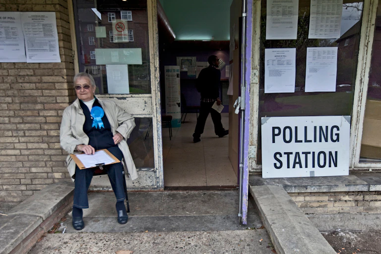 A Conservative party representative sitting on a chair outside a polling station.
