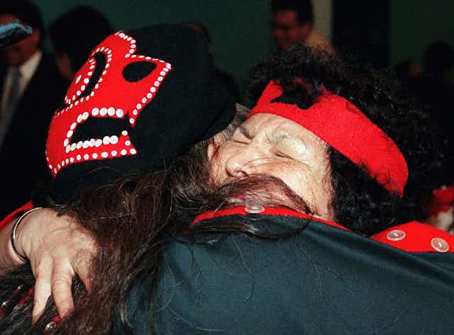 Two people wearing Indigenous clothing hug.