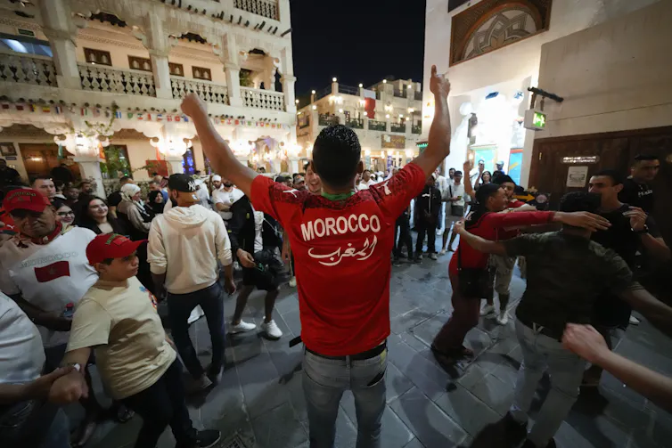 Un homme de dos, arborant un chandail où il est écrit ‘Morocco », salue une foule