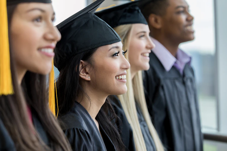 Four college students wearing graduation caps and gowns look onward to the right.