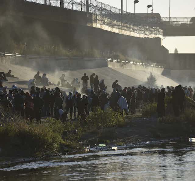 Hundreds of people are standing on a river bank near a concrete wall that is topped with a metal fence.