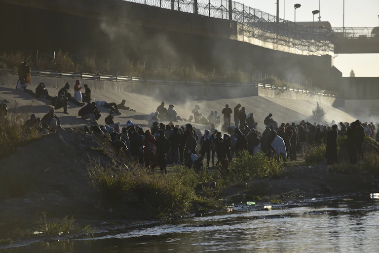 Hundreds of people are standing on a river bank near a concrete wall that is topped with a metal fence.