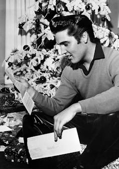 A black and white photograph of a man with slicked-back hair in a sweater kneeling by a Christmas tree as he reads a letter.