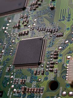 A computer chip with a black square surrounded by metal dots and lines on a green board.