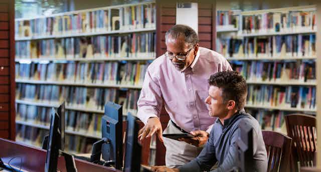 An older man points things out to a middle aged man while they look at computer screens in a library.