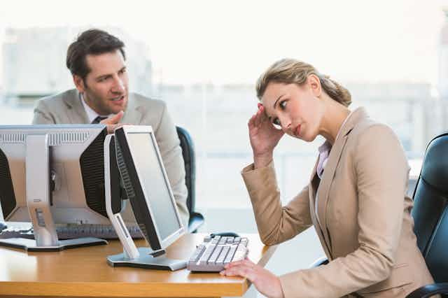 A woman in business attire looks at her computer screen while avoiding the eyes of her male colleague, who is gesturing and speaking to her