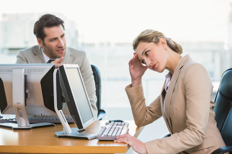 A woman in business attire looks at her computer screen while avoiding the eyes of her male colleague, who is gesturing and speaking to her