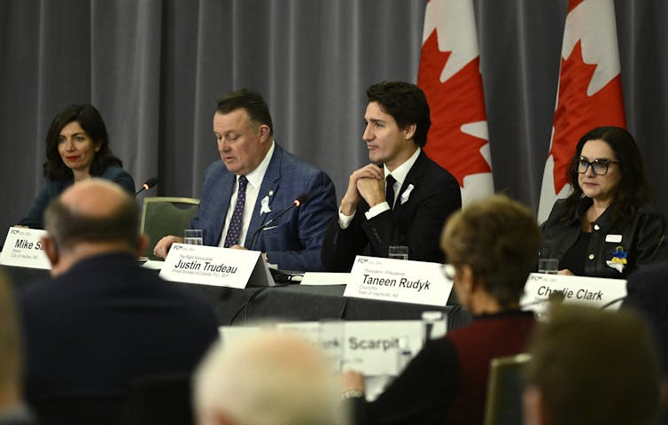A dark-haired man sits among men and women at a round table, Canadian flags behind them.
