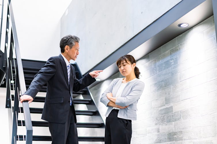 An older man in a business suit points and talks sternly to a younger woman who is also in business attire, depicting mansplaining
