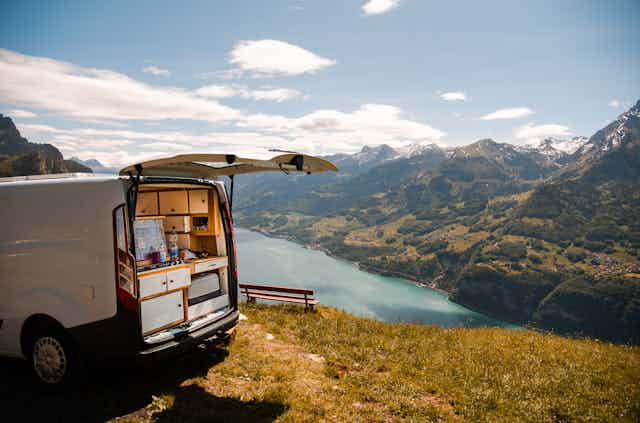 A van customized for living sits atop a ridge overlooking a river with mountains in the background
