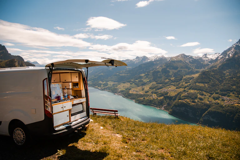 A van customized for living sits atop a ridge overlooking a river with mountains in the background