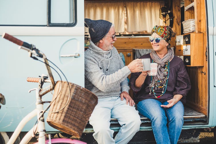 An older adult couple sit on the side of their van and clink their coffee mugs together
