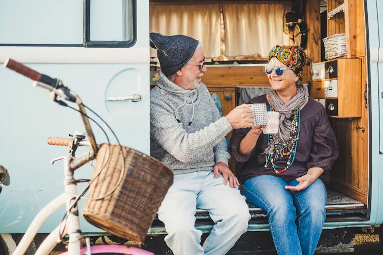 An older adult couple sit on the side of their van and clink their coffee mugs together