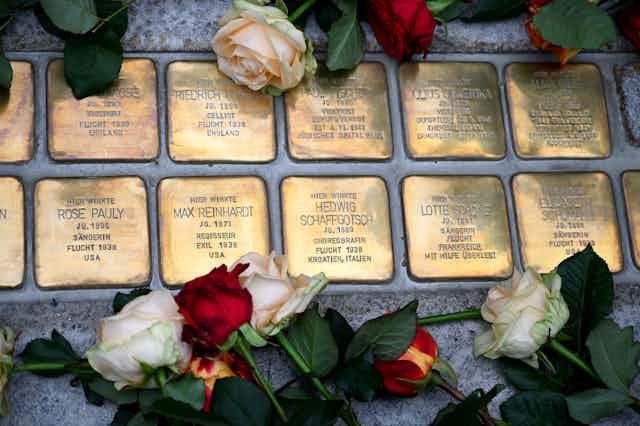 10 small bronze plaques with people's names on them are seen placed in cement, with white and red roses surrounding them.