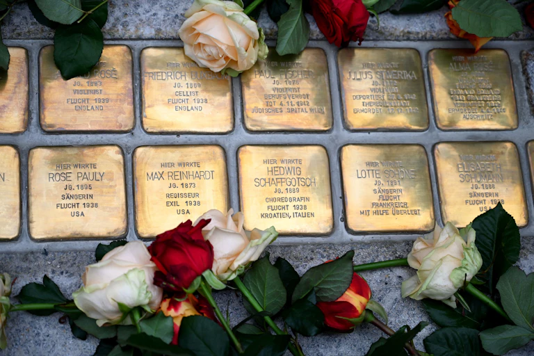10 small bronze plaques with people's names on them are seen placed in cement, with white and red roses surrounding them.