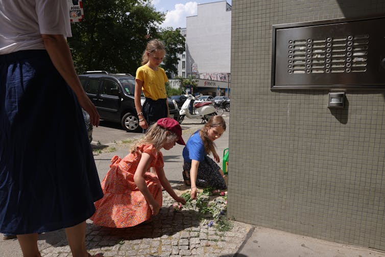 Three school aged blond girls sit and stand over cobblestones on a sidewalk and appear to place flowers there.