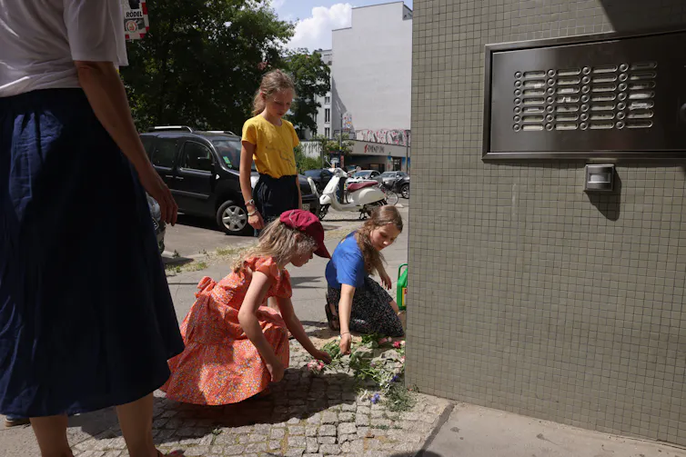 Three school aged blond girls sit and stand over cobblestones on a sidewalk and appear to place flowers there.