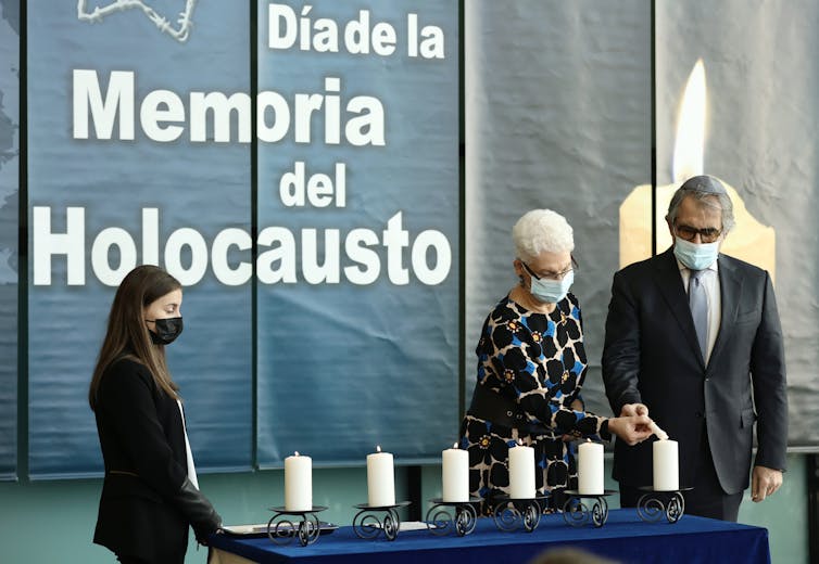 Three people stand near white candles on a table, in front of a banner that says 'dia de la memoria del Holocausto' behind them.