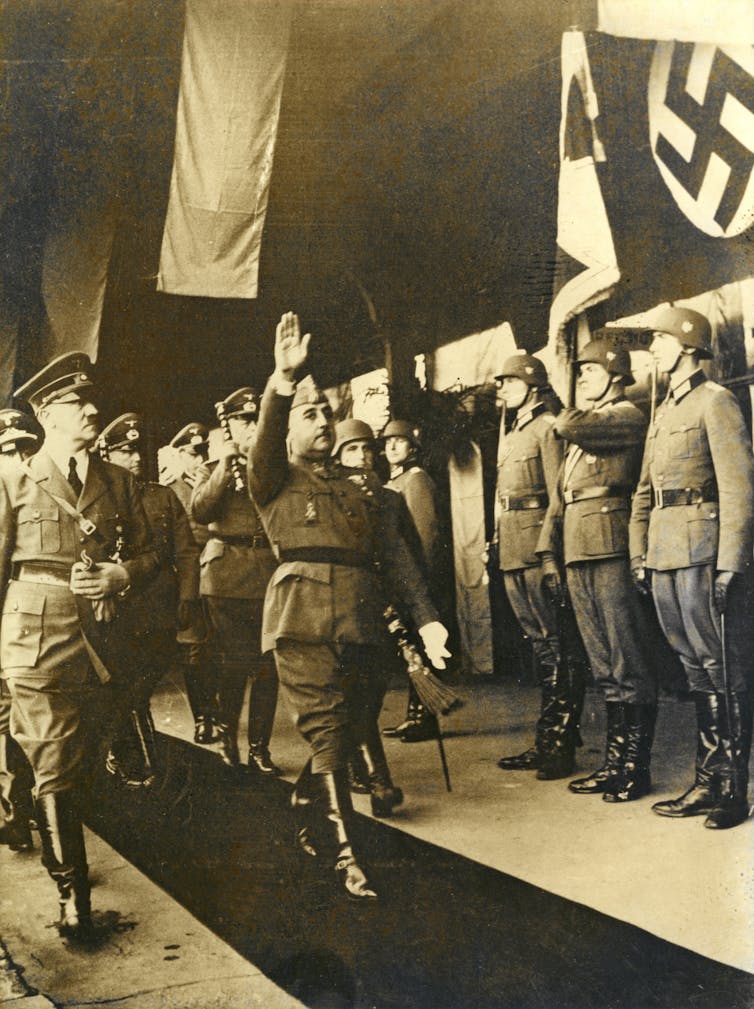 A black and white photo shows two men in military clothing, with one doing a heil salute, next to a row of soldiers, some of whom hold a Nazi flag