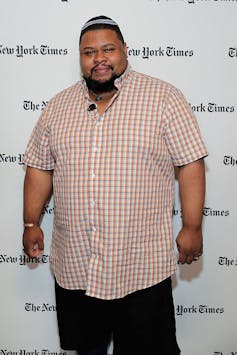 A Black man wearing a white and red checked shirt and black pants stands in front of a backdrop with the words The New York Times