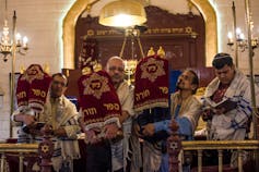 A handful of men hold scrolls with Hebrew lettering at the front of a synagogue.