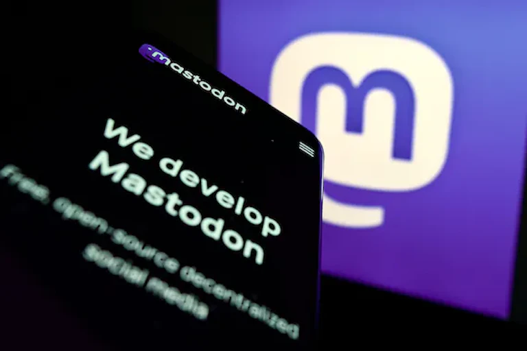 logo mastodon tampil di sebuah layar — huruf m biru dalam gelembung percakapan putih — ada layar ponsel yang menampilkan teks WE DEVELOP MASTODON