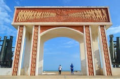 The Door of No Return in Ouidah. A large archway facing the sea. It is cream and bronze in colour with stylised bas-relief sculptures on it showing chained human bodies and ships | jbdodane/Wikimedia Commons, CC BY - 