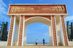 A large archway facing the sea. It is cream and bronze in colour with stylised bas-relief sculptures on it showing chained human bodies and ships.