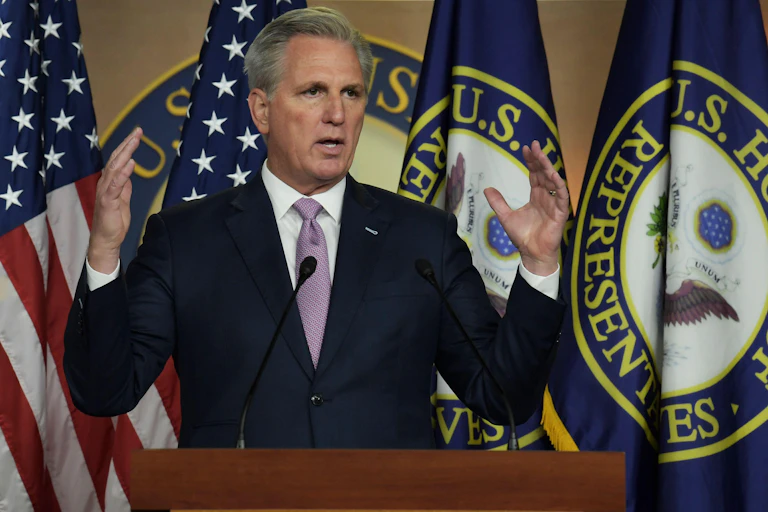 A man in a suit standing in front of the US flag.