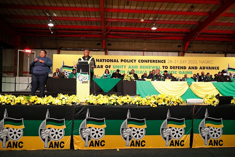 A woman wearing pants uses sign language to interpretes a speech by a man wearing a leather jacket speaking at a podium to her left.