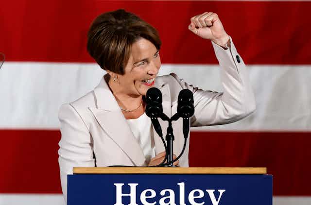 A happy, smiling woman with dark hair and in a white jacket at a lectern, raising her fist in triumph.