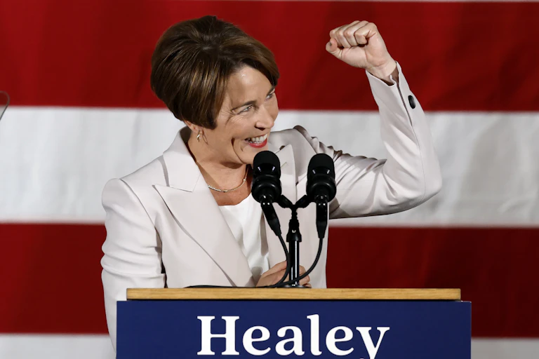 A happy, smiling woman with dark hair and in a white jacket at a lectern, raising her fist in triumph.