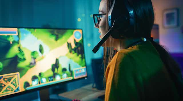 A girl with glasses and a pony tail plays a video game on a computer screen, wearing a large headset. The screen shows a map of a forest area. 