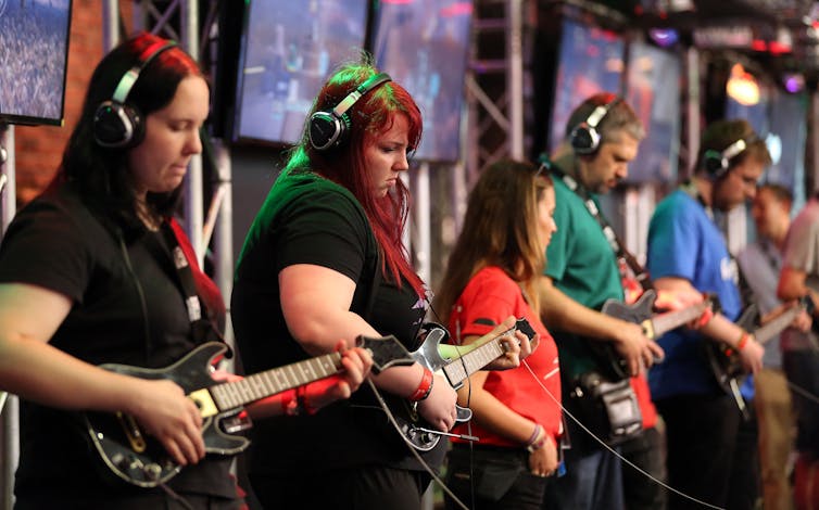 Line of women and men playing guitars with headphones on in front of screens