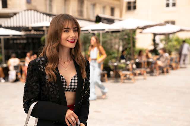 Emily stands in a Parisian courtyard wearing a blazer over a gingham bra. 